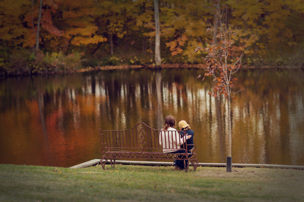 A teenage girl and her younger brother sit on a metal bench looking out at the lake with the gorgeous fall colors.