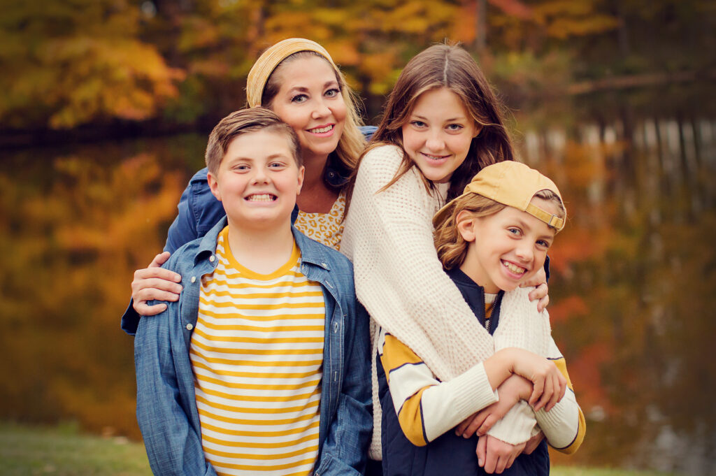 A family in fall colors smiles at the camera with a beautiful Indiana fall backdrop behind them.