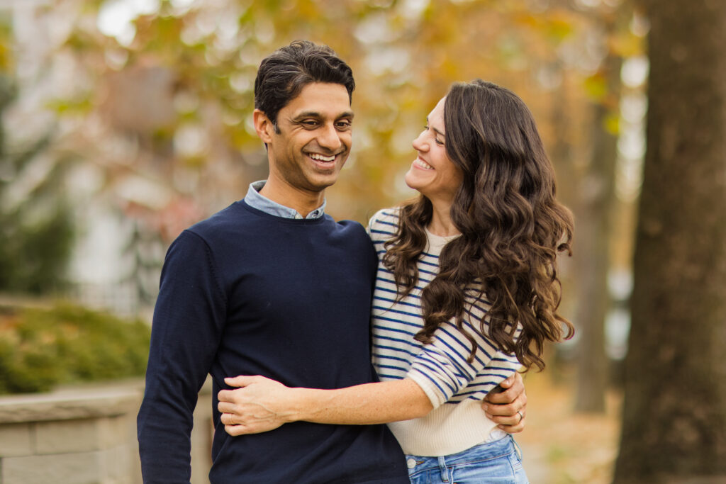 Man in blue sweater and woman in blue and white striped sweater hugging and smiling with fall leaves in the background.