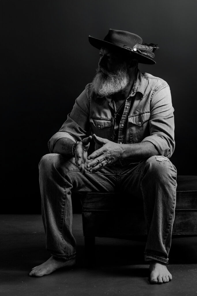 Man in denim shirt and jeans and hat with feathers sitting on stool looking off to side with dramatic lighting.
